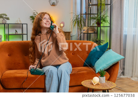 Smiling young woman making wired telephone call conversation with friends sitting on sofa at home Smiling young woman making wired telephone call conversation with friends sitting on sofa at home 125677038