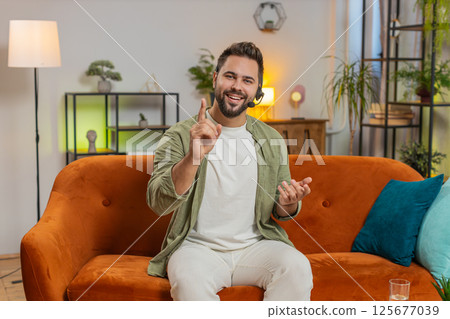 Confident young Caucasian man guy sitting on sofa at home wearing headset talking with client 125677039