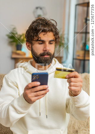 Happy young Hispanic man guy using credit bank card and cellphone while shopping online at home 125677356
