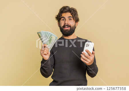 Successful Lebanese young man using smartphone and holding money banknotes against beige background Successful Lebanese young man using smartphone and holding money banknotes against beige background 125677358