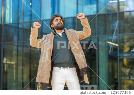 Excited man looking surprised clenching fists, shocked by sudden victory standing on city street 125677366