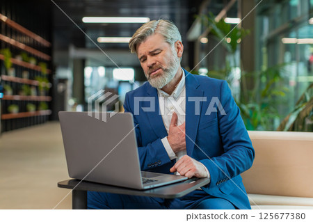 Mature businessman using laptop and suffers from tension holding chest with heart attack in lobby 125677380