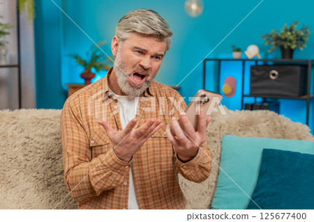 Mature man having phone argument on speaker, expressing stress and negative emotions on sofa at home Mature man having phone argument on speaker, expressing stress and negative emotions on sofa at home 125677400