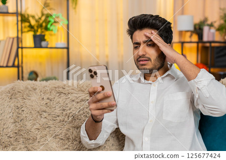 Indian man upset looking at smartphone, hand on forehead, reading bad news, feeling worried 125677424