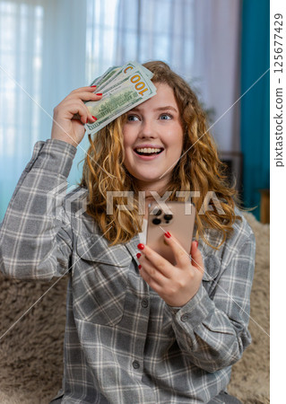 Young woman sits on sofa smiling happily showing thumbs up after receiving money SMS on smartphone Young woman sits on sofa smiling happily showing thumbs up after receiving money SMS on smartphone 125677429