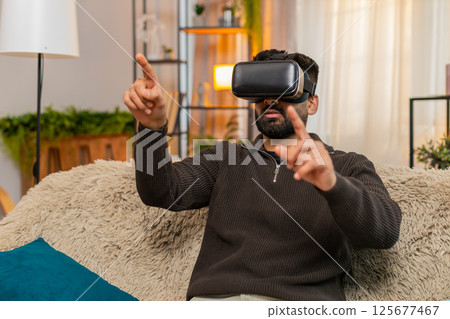 Indian man on sofa wears VR headset at home and reacts with excitement to immersive virtual world Indian man on sofa wears VR headset at home and reacts with excitement to immersive virtual world 125677467