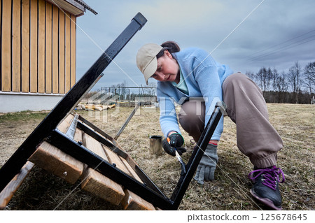 Painting metal frame of garden bench with brush, woman applies protective coating to prevent rust and extend bench lifespan outdoors near wooden house. 125678645