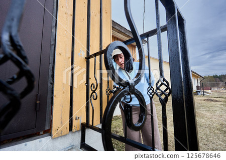 Standing at entrance to country house woman carefully paints porch railing transforming metal bars into glossy black surface against rural background. Standing at entrance to country house woman carefully paints porch railing transforming metal bars into glossy black surface against rural background. 125678646