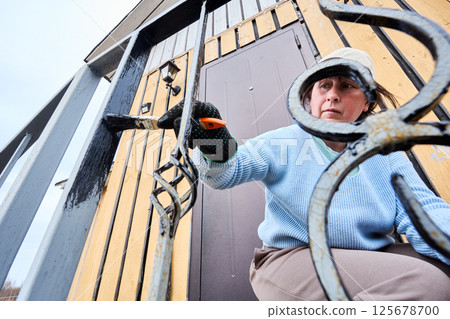 Mature Caucasian woman applies black metal paint to porch railings at entrance to country house. 125678700