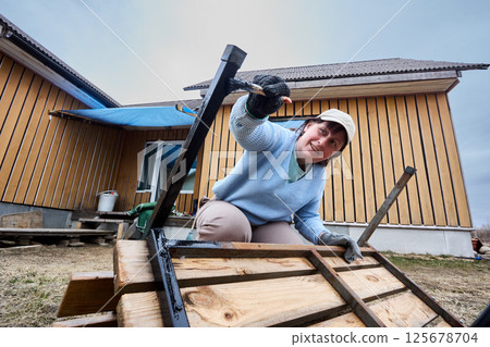 Painting metal parts of garden bench with brush, woman applies black paint carefully outdoors near wooden houses under cloudy sky. 125678704