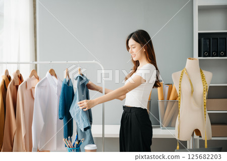 Successful fashion designer. Attractive young asian woman with smile while standing in workshop. Beautiful owner business woman working and holds tablet, laptop and smartphone in studio. 125682203