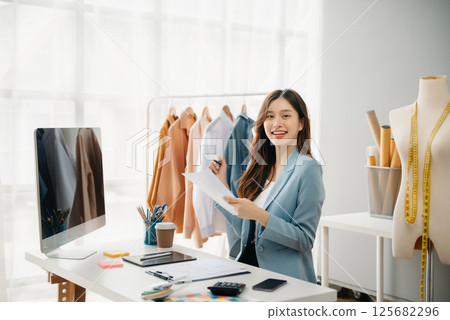 Fashion designer woman talking smart phone and using laptop with digital tablet computer in modern studio the clothes hanging on the racks 125682296