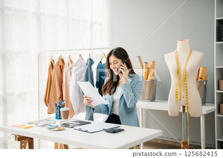 Fashion designer woman talking smart phone and using laptop with digital tablet computer in modern studio the clothes hanging on the racks Fashion designer woman talking smart phone and using laptop with digital tablet computer in modern studio the clothes hanging on the racks 125682415