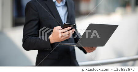 Businesswoman s hands typing on smartphone and laptop keyboard in morning light computer, typing, online in outside office Businesswoman s hands typing on smartphone and laptop keyboard in morning light computer, typing, online in outside office 125682673