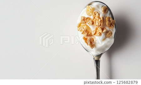 A spoon filled with creamy yogurt and crunchy cereal flakes rests on a clean white background 125682879