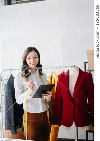 Diverse female fashion designers at work with tailor centimeters on necks and holds tablet and smartphone. Diverse female fashion designers at work with tailor centimeters on necks and holds tablet and smartphone. 125683869