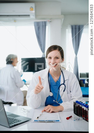 Confident young Caucasian female doctor in white medical uniform sit at desk working on computer. Smiling use laptop write in medical journal 125684383