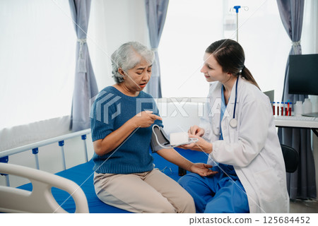 Doctor in white suit take notes while discussing and Asian elderly, woman patient who lying on bed with receiving saline solution in hospital or clinic. Doctor in white suit take notes while discussing and Asian elderly, woman patient who lying on bed with receiving saline solution in hospital or clinic. 125684452