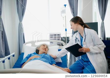 Doctor in white suit take notes while discussing and Asian elderly, woman patient who lying on bed with receiving saline solution in hospital or clinic. 125684454