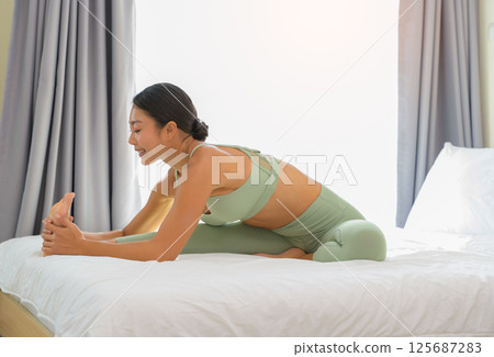 Woman in green activewear practicing yoga on bed in morning. Asian female stretching legs with forward bend in bedroom at home Woman in green activewear practicing yoga on bed in morning. Asian female stretching legs with forward bend in bedroom at home 125687283
