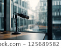 A professional podcast microphone is mounted on a stand atop a wooden desk. The desk is situated beside a large window in a modern office building, allowing natural light to fill the minimalist 125689582