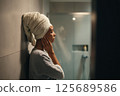 A Black woman, around 29 years old, stands against a tiled bathroom wall with a towel around her shoulders. Her eyes are closed and her expression is peaceful, enjoying a moment of relaxation and self 125689586