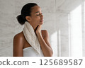 A young Black woman, approximately 28 years old, stands with a towel draped over her shoulders and wipes her neck. Her eyes are closed and she appears relaxed, surrounded by soft natural light and a 125689587
