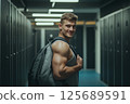 A fit young white male, around 25 years old, stands in a gym locker room wearing a tank top and holding a backpack over one shoulder. He smiles confidently, surrounded by rows of dark lockers and 125689591