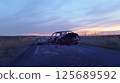 A single severely wrecked car sits deserted on a narrow, winding country road during dusk. The surrounding landscape is flat and empty, with tall grass lining the road and a wide, overcast sky 125689592