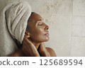 A young Black woman, around 30 years old, is seen in a serene moment with a towel wrapped around her head. Her eyes are closed and her skin glows with moisture as she leans gently against a light 125689594
