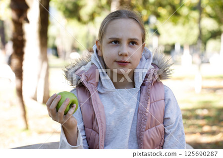 Playful kid holding green apple. Young girl in park, outdoors portrait 125690287