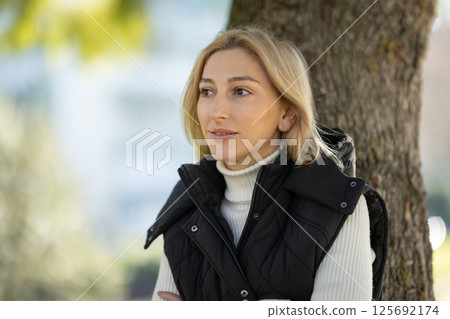 Blond woman leaning against tree enjoying day in park 125692174