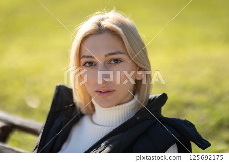 Portrait of a blonde woman sitting on a bench, enjoying a sunny day in a park 125692175