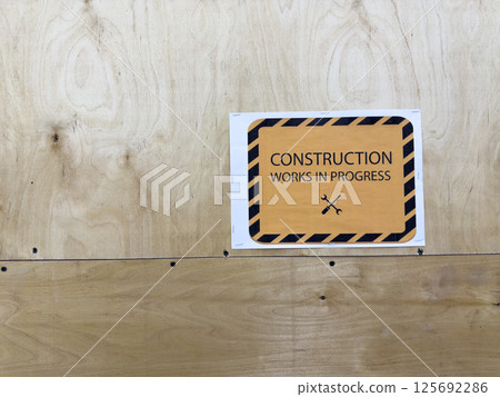 Warning sign with text construction works in progress attached to plywood wall, safety notice at building site, caution board with tools icon indicating maintenance or renovation zone Warning sign with text construction works in progress attached to plywood wall, safety notice at building site, caution board with tools icon indicating maintenance or renovation zone 125692286