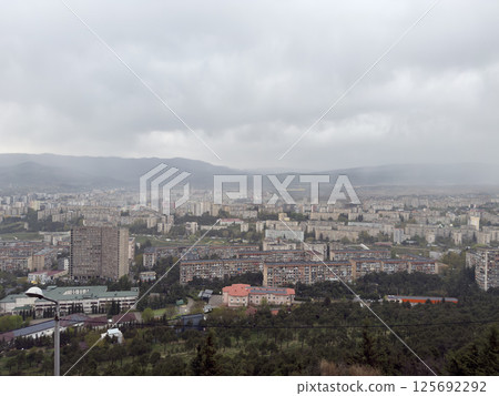 Overcast cityscape with dense apartment blocks and distant hills under cloudy sky. Urban skyline on rainy day showing architecture, density and atmosphere of modern residential development Overcast cityscape with dense apartment blocks and distant hills under cloudy sky. Urban skyline on rainy day showing architecture, density and atmosphere of modern residential development 125692292