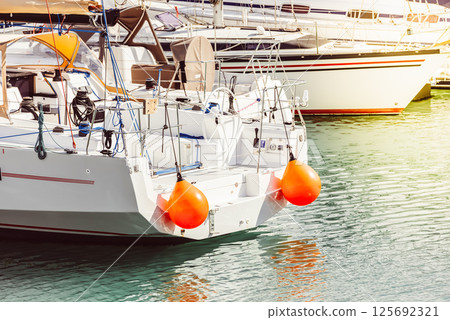 Yacht with orange buoys docked at marina, calm sea reflecting light Yacht with orange buoys docked at marina, calm sea reflecting light 125692321
