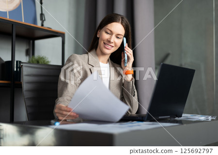 Business female talking on phone at desk and holding a document. Business female talking on phone at desk and holding a document. 125692797