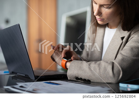 Young female professional is sitting at a desk in an office using smartwatch on her wrist. Young female professional is sitting at a desk in an office using smartwatch on her wrist. 125692804