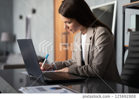 Young woman sitting at desk in office, working on her laptop and writing notes documents home office 125692808