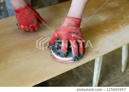 Close-up view of a carpenter wearing red protective gloves sanding a wooden board using a manual sanding tool 125693957