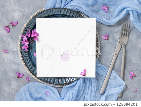 Wedding table place with an envelope near fabric and wild purple flowers top view on gray, mockup 125694201