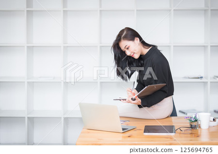 Asian businesswoman sitting on the edge of desk and working in modern office. Asian businesswoman sitting on the edge of desk and working in modern office. 125694765