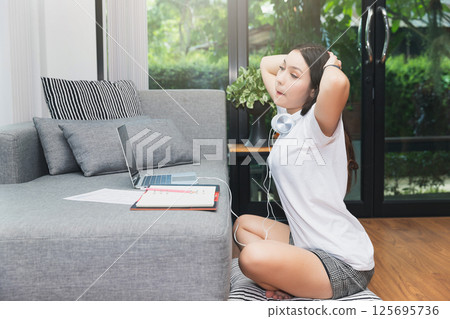 A woman with headphones siting and stretching lazily or stretching herself after using her laptop for a long time on the living room floor. A woman with headphones siting and stretching lazily or stretching herself after using her laptop for a long time on the living room floor. 125695736