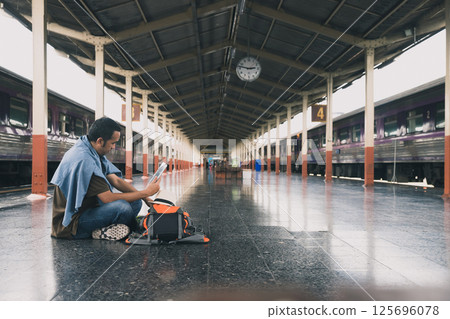 A young man traveler with digital tablet sitting on floor at train station choose where to travel. Concept of backpackers travel adventure by train. 125696078
