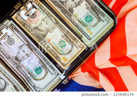 Dollars cash with U.S. filled briefcase resting an American flag symbolizing wealth patriotism in business setting Dollars cash with U.S. filled briefcase resting an American flag symbolizing wealth patriotism in business setting 125696154
