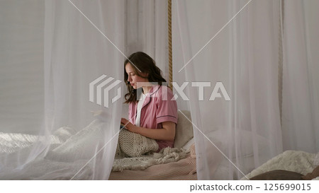 A young woman sits in a serene bedroom surrounded by white curtains, deeply focused on a book. This image highlights the importance of finding tranquility in a busy, modern world. A young woman sits in a serene bedroom surrounded by white curtains, deeply focused on a book. This image highlights the importance of finding tranquility in a busy, modern world. 125699015