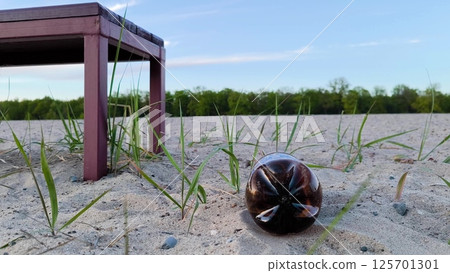 A brown plastic bottle is lying like garbage on the beach, next to a bench for relaxing. A plastic bottle is like trash on the beach. Close up pedestal shot 125701301