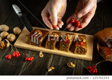 A baker arranges four slices of moist cake on a wooden platter, adding red berries and walnuts as a garnish. The rustic kitchen features dark wood and natural lighting 125701355