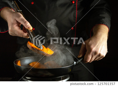 A chef expertly uses tongs to place sweet potato slices into a frying pan on the stove. Steam rises, emphasizing the cooking process in a dimly lit kitchen A chef expertly uses tongs to place sweet potato slices into a frying pan on the stove. Steam rises, emphasizing the cooking process in a dimly lit kitchen 125701357