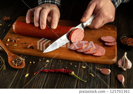 A person slices a sausage on a wooden board, surrounded by spices, garlic, and herbs. The warm evening light highlights the rustic kitchen atmosphere, enhancing the culinary experience 125701361